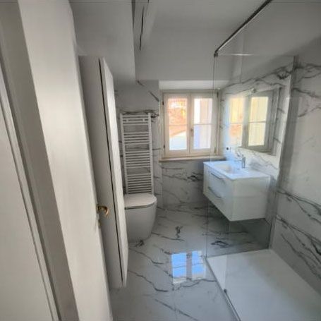 Bagno moderno con piastrelle effetto marmo bianco, doccia in vetro, lavabo a parete e WC bianco vicino a una finestra.