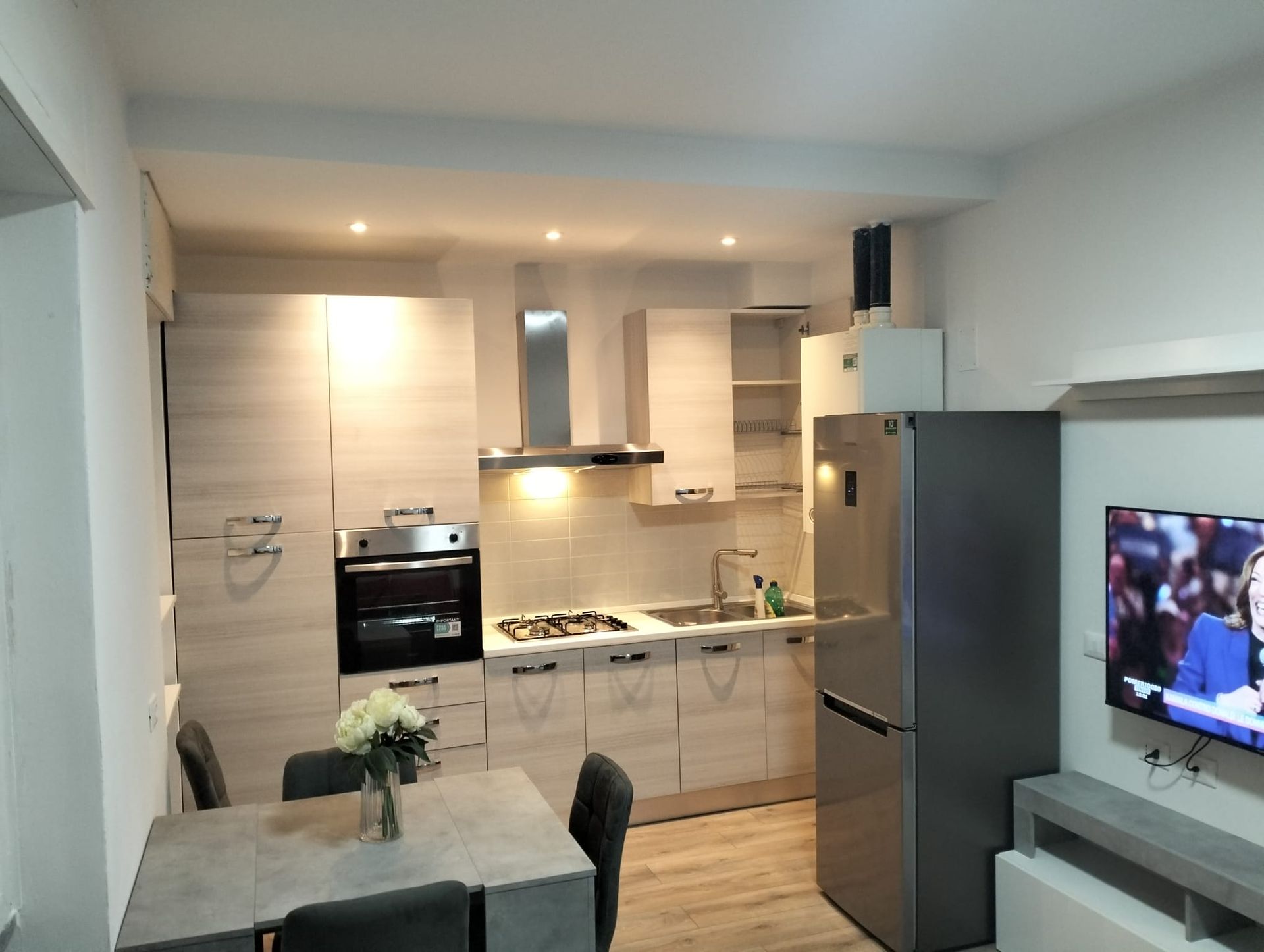 Una cucina moderna con mobili in legno chiaro, elettrodomestici in acciaio inossidabile e un tavolo da pranzo grigio accanto a un televisore a parete.