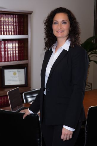 A woman in a suit and white shirt is standing in front of a bookshelf.