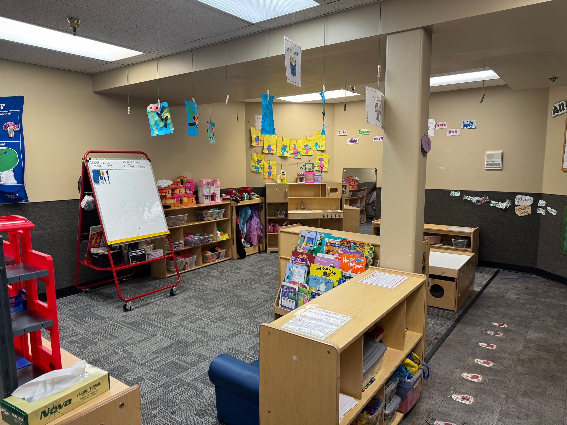 A classroom with bookshelves, toys, and a whiteboard. Artwork hangs from the ceiling.
