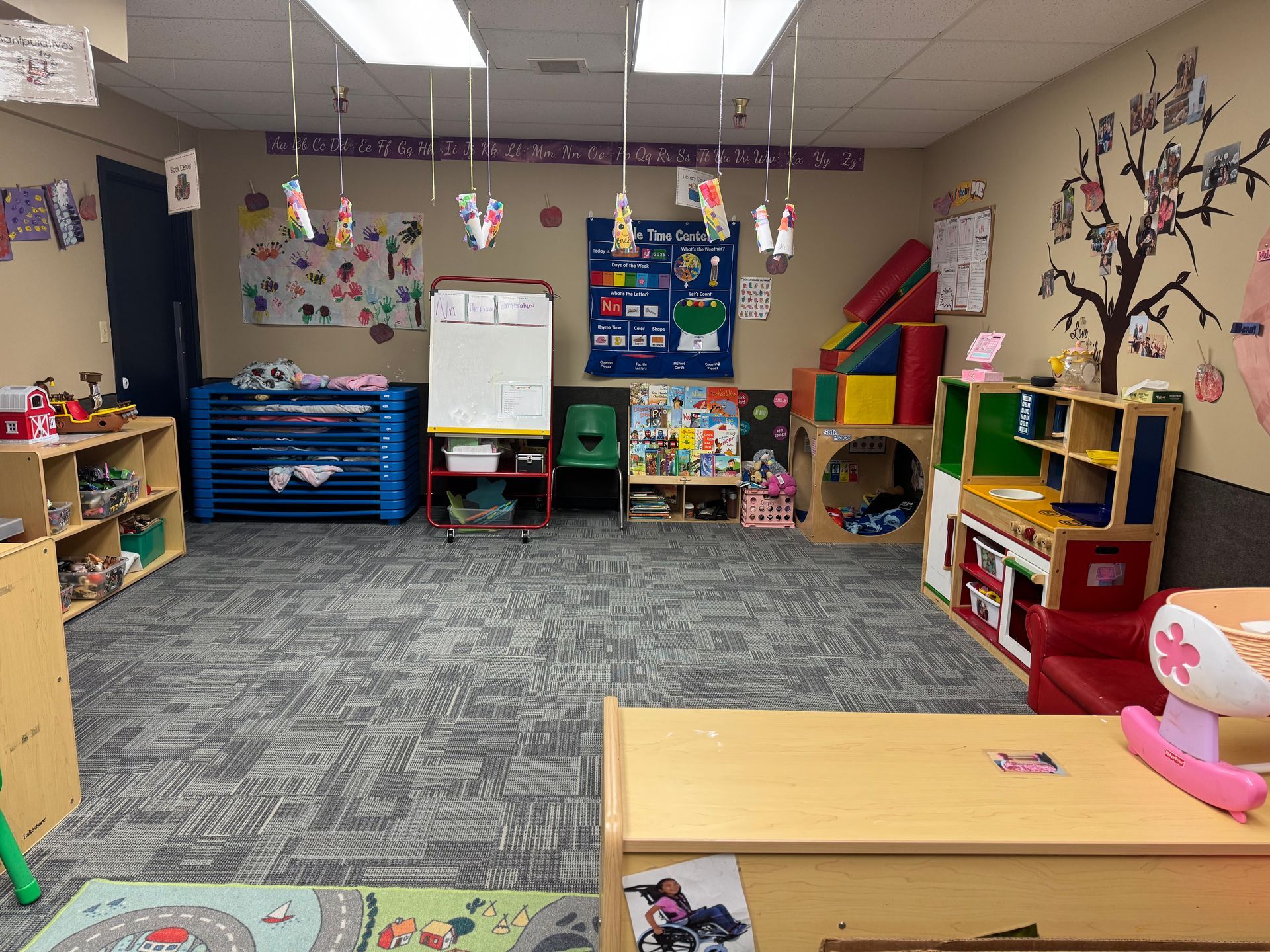 A brightly lit preschool classroom. Toys, artwork, and play structures are arranged throughout the room.