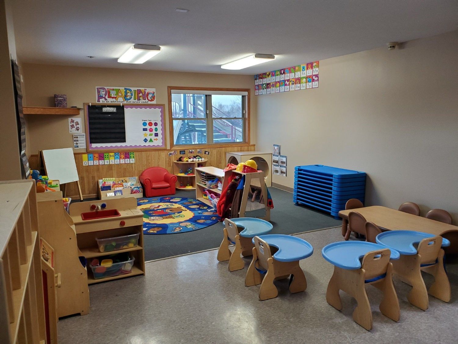 Childcare room, light brown walls, small blue tables, toys, window, and low wooden shelves.