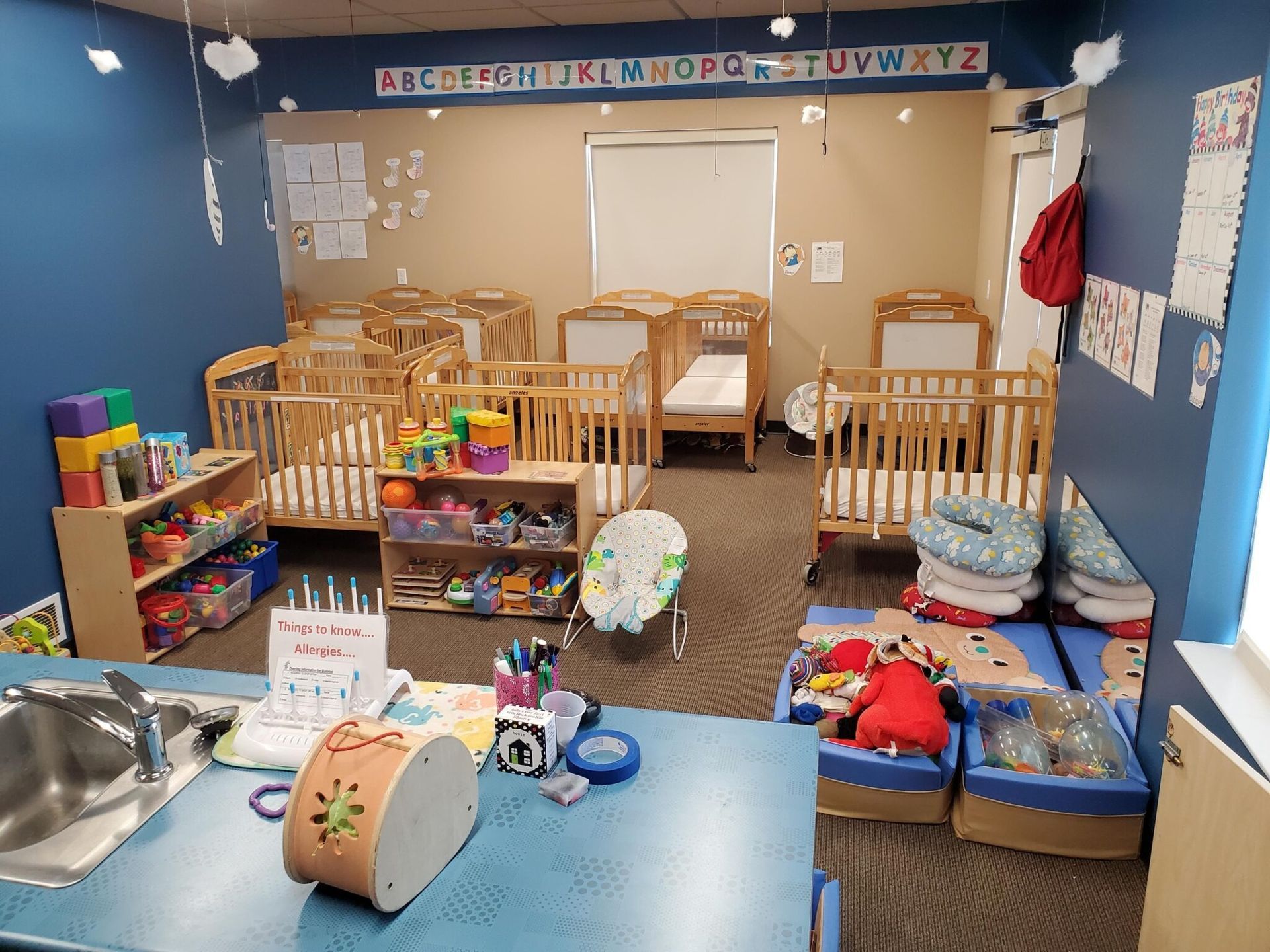 Daycare room with cribs, toys, and a sink; blue and tan walls.
