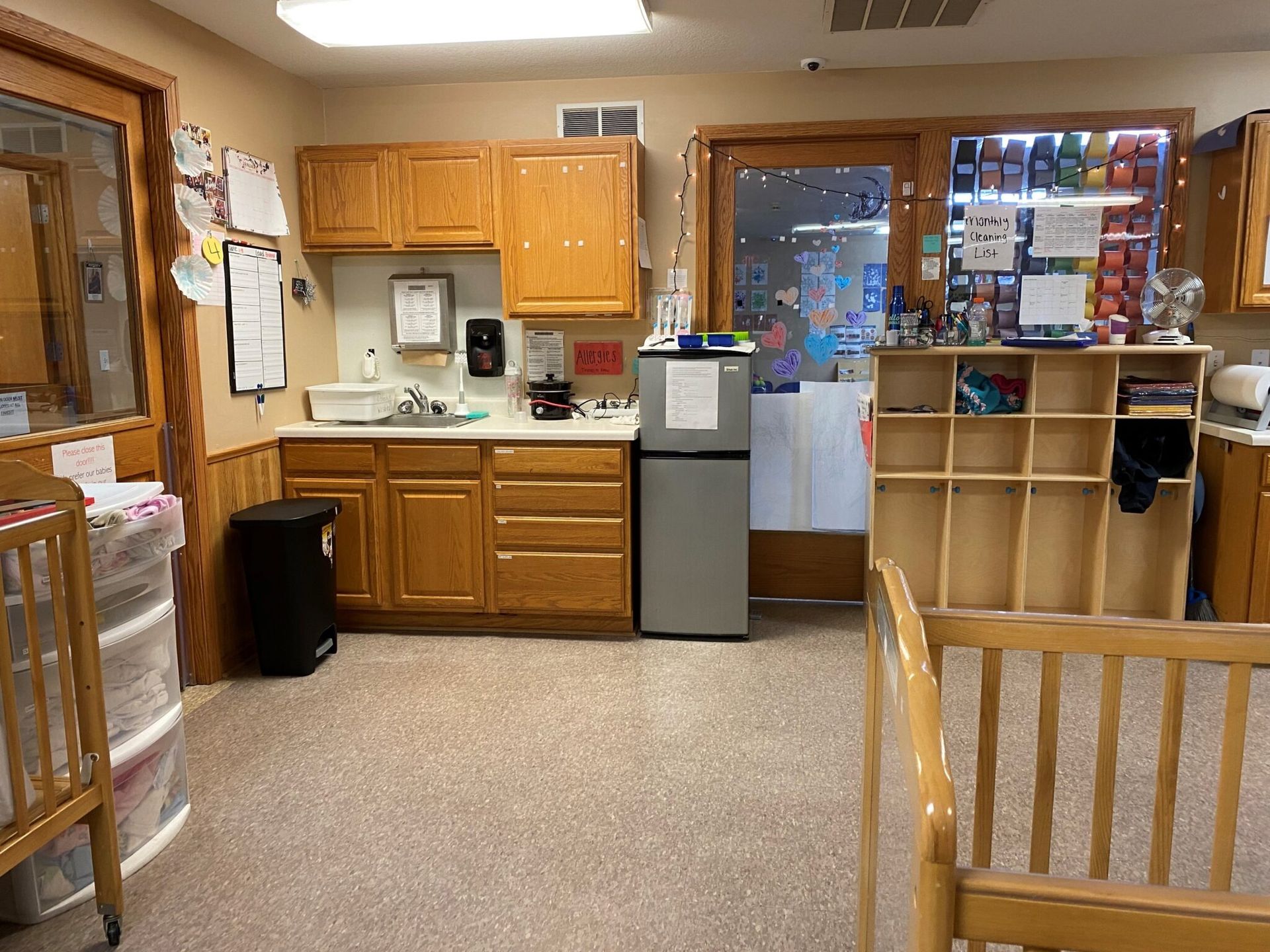 Daycare kitchen with cabinets, sink, refrigerator, and cubby shelves. Two cribs are in view.
