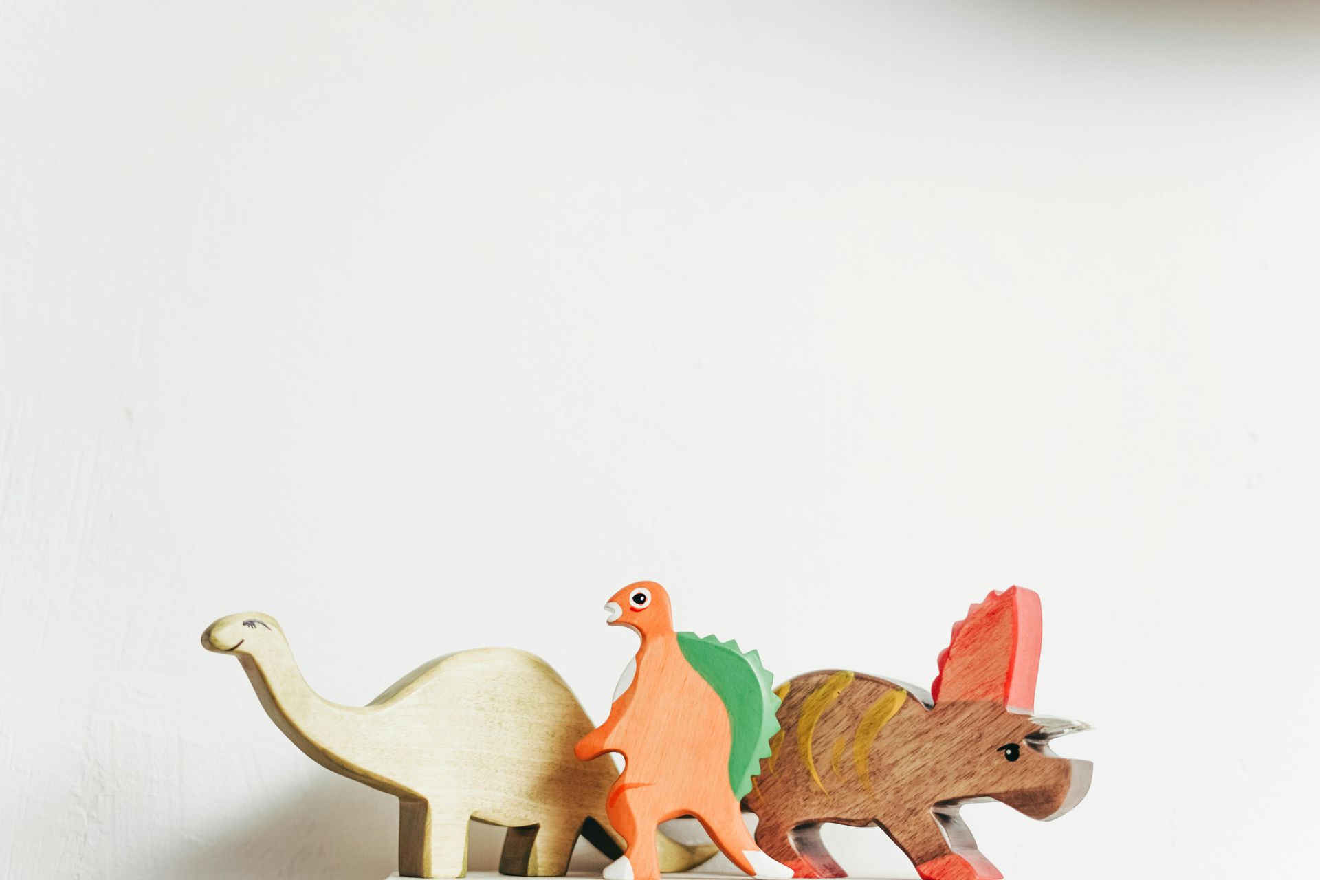 Wooden dinosaur toys: tan brontosaurus, orange stegosaurus, and brown triceratops against a white background.