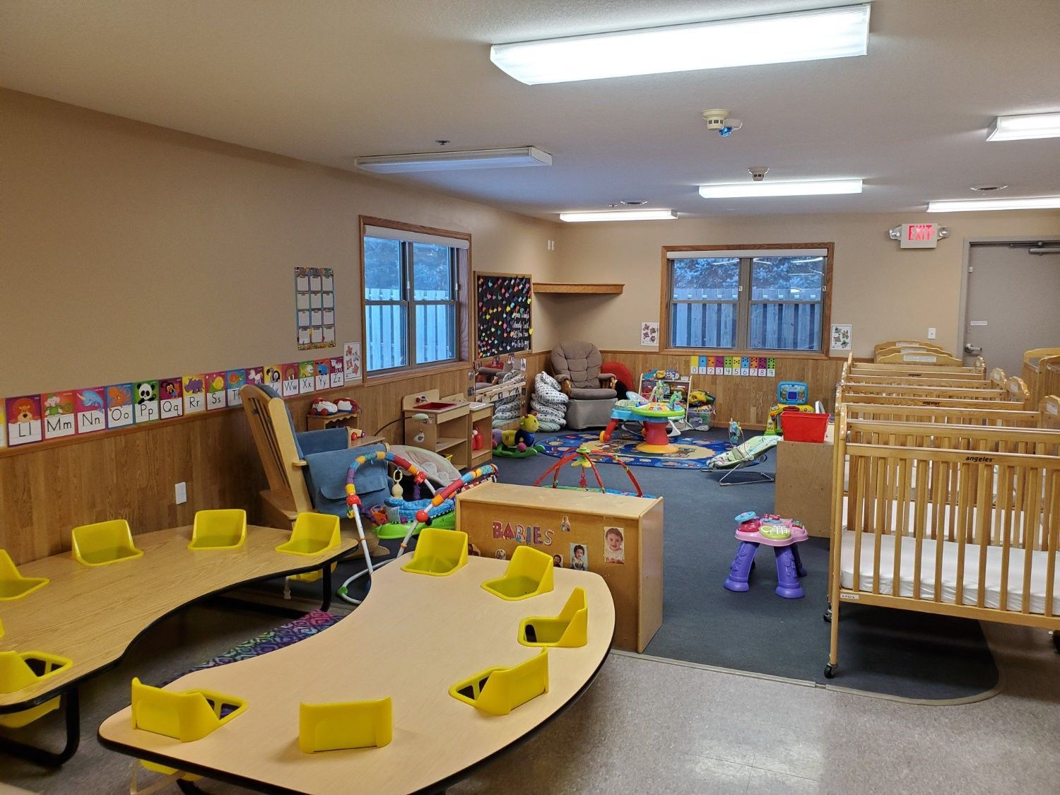 Childcare room with tables, chairs, toys, cribs, and windows.
