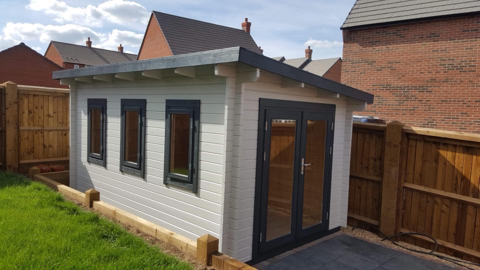 A small white building with a roof and windows is in a backyard next to a wooden fence.