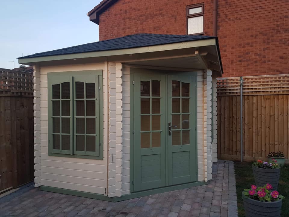 A small white building with green doors and windows in front of a brick house.