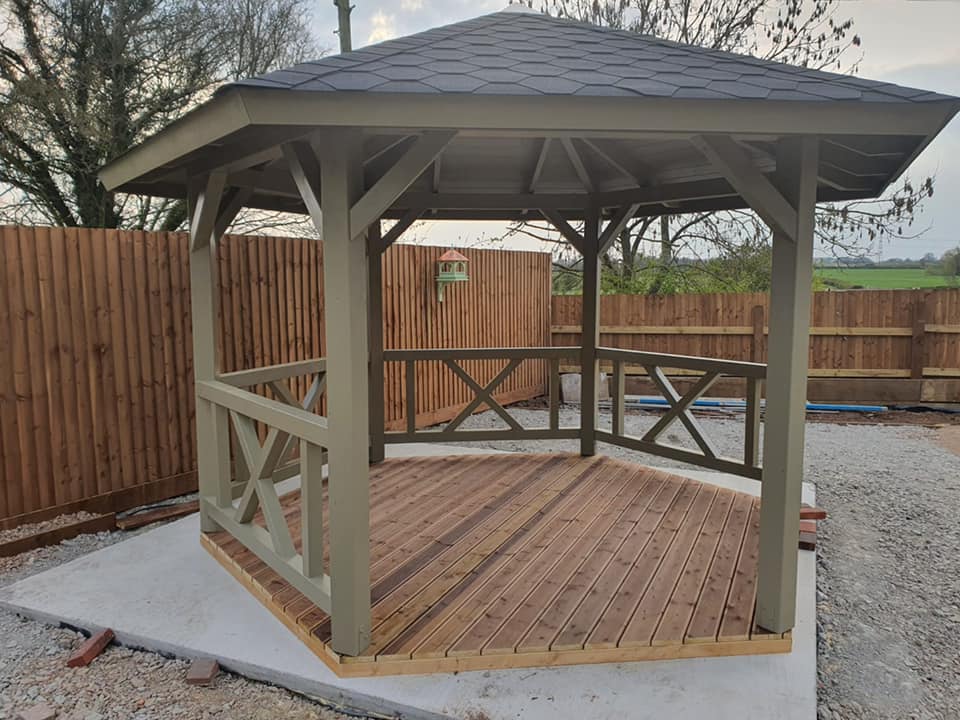 A wooden gazebo with a slate roof is sitting in front of a wooden fence.