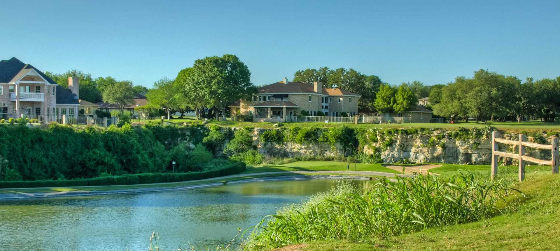 There is a lake in the middle of a field with houses in the background.
