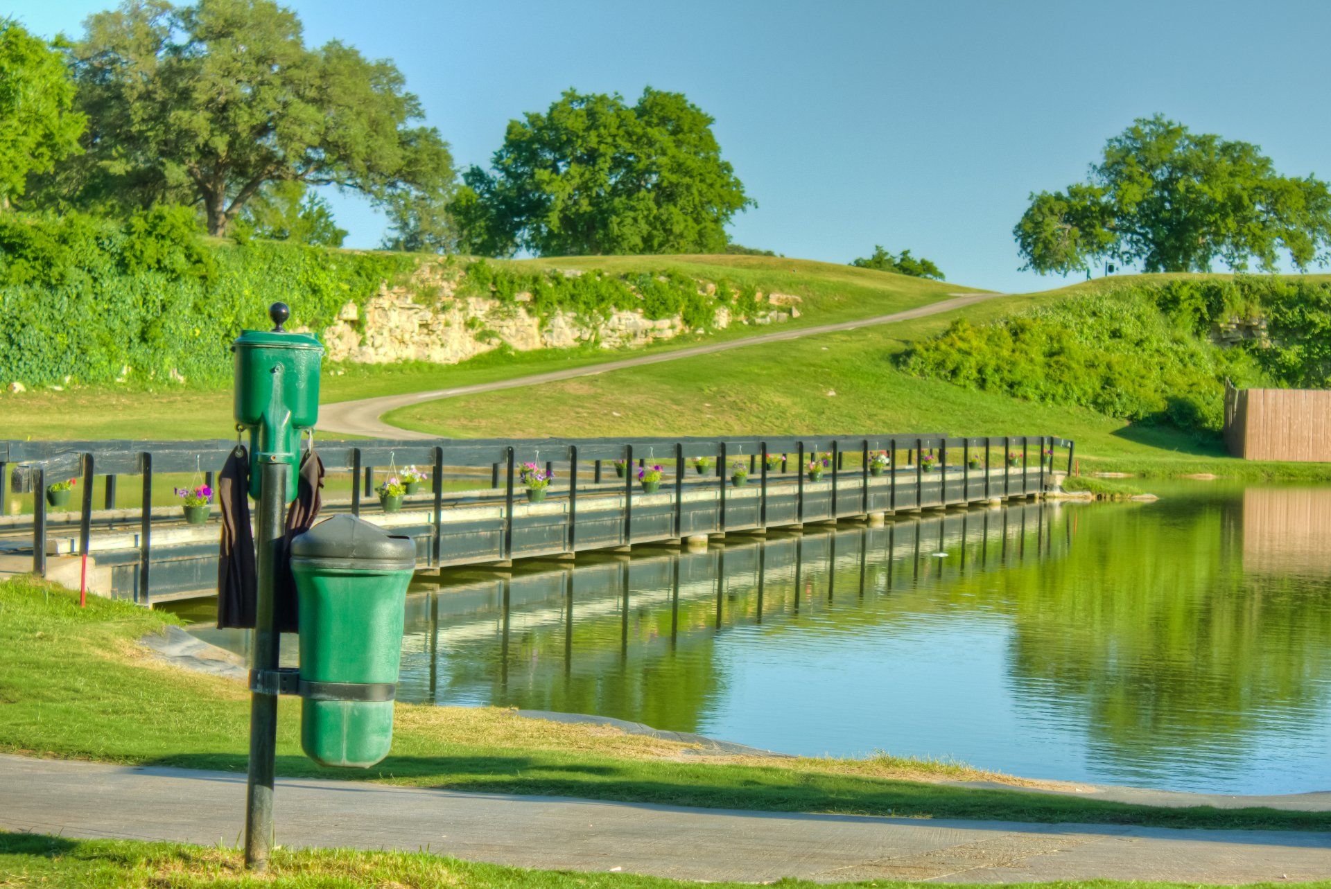A green trash can is sitting next to a pond in a park.