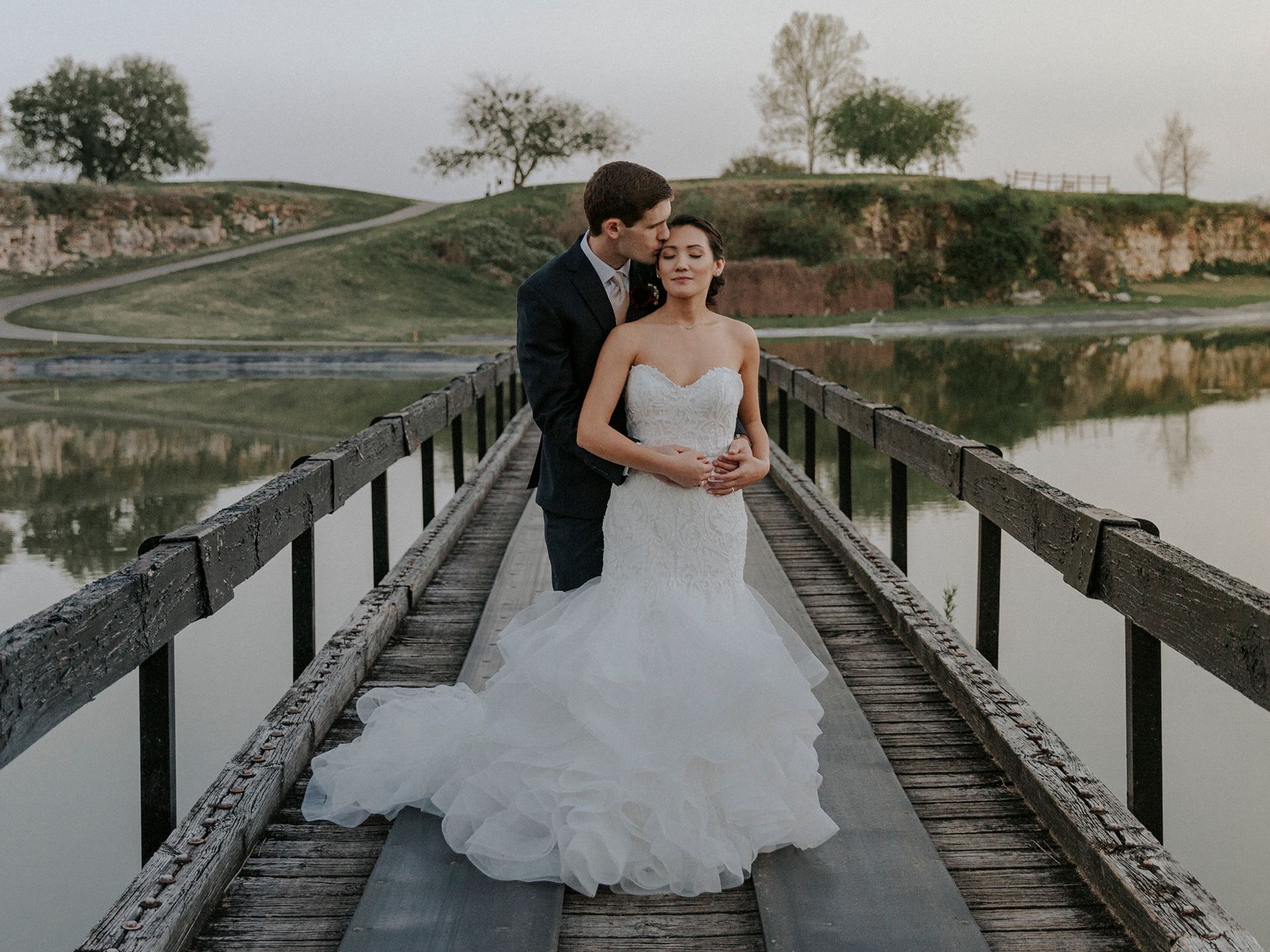 A bride and groom are posing for a picture on a wooden bridge over a lake.