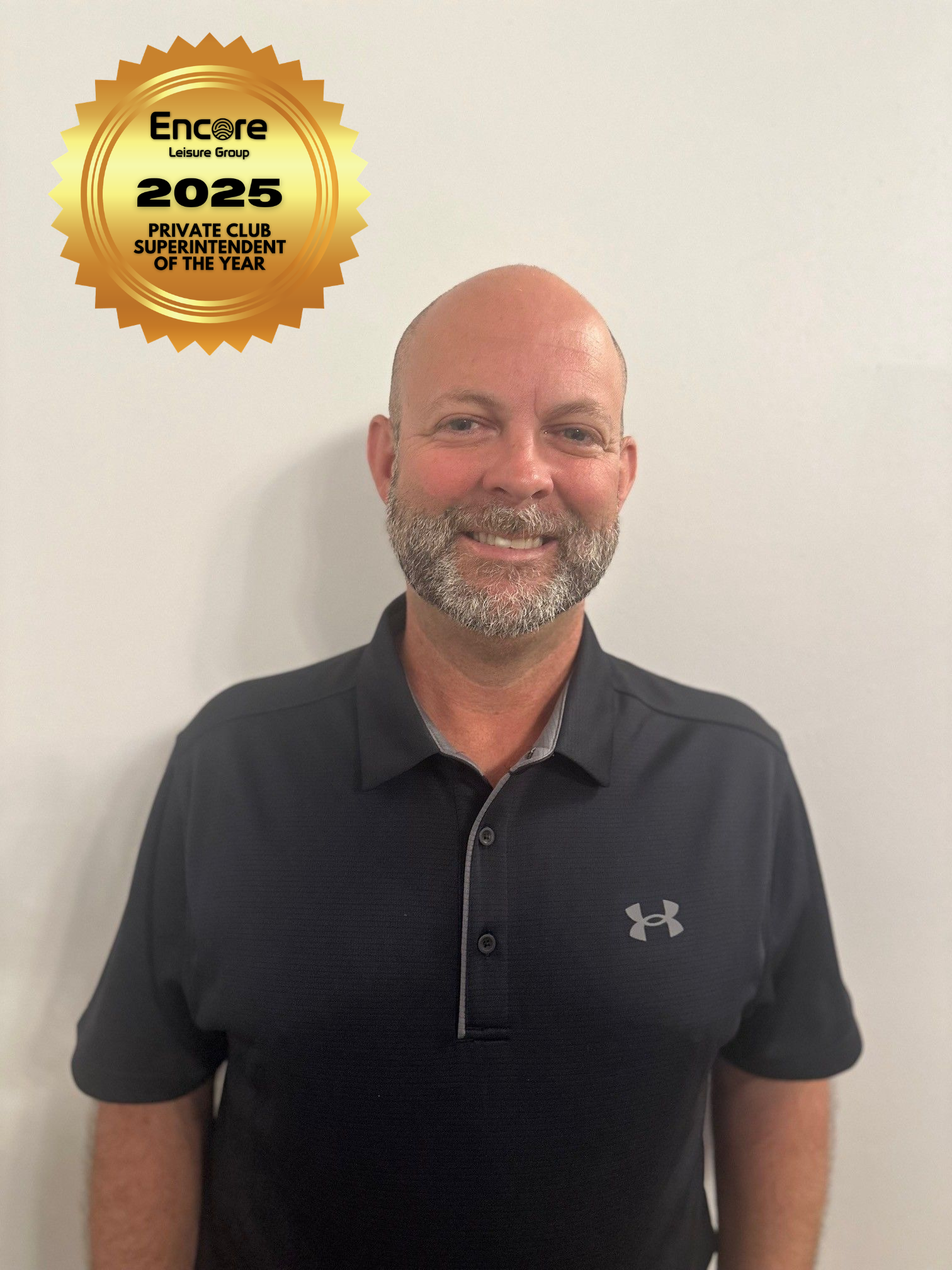 A bald man with a beard is wearing a black under armour polo shirt.