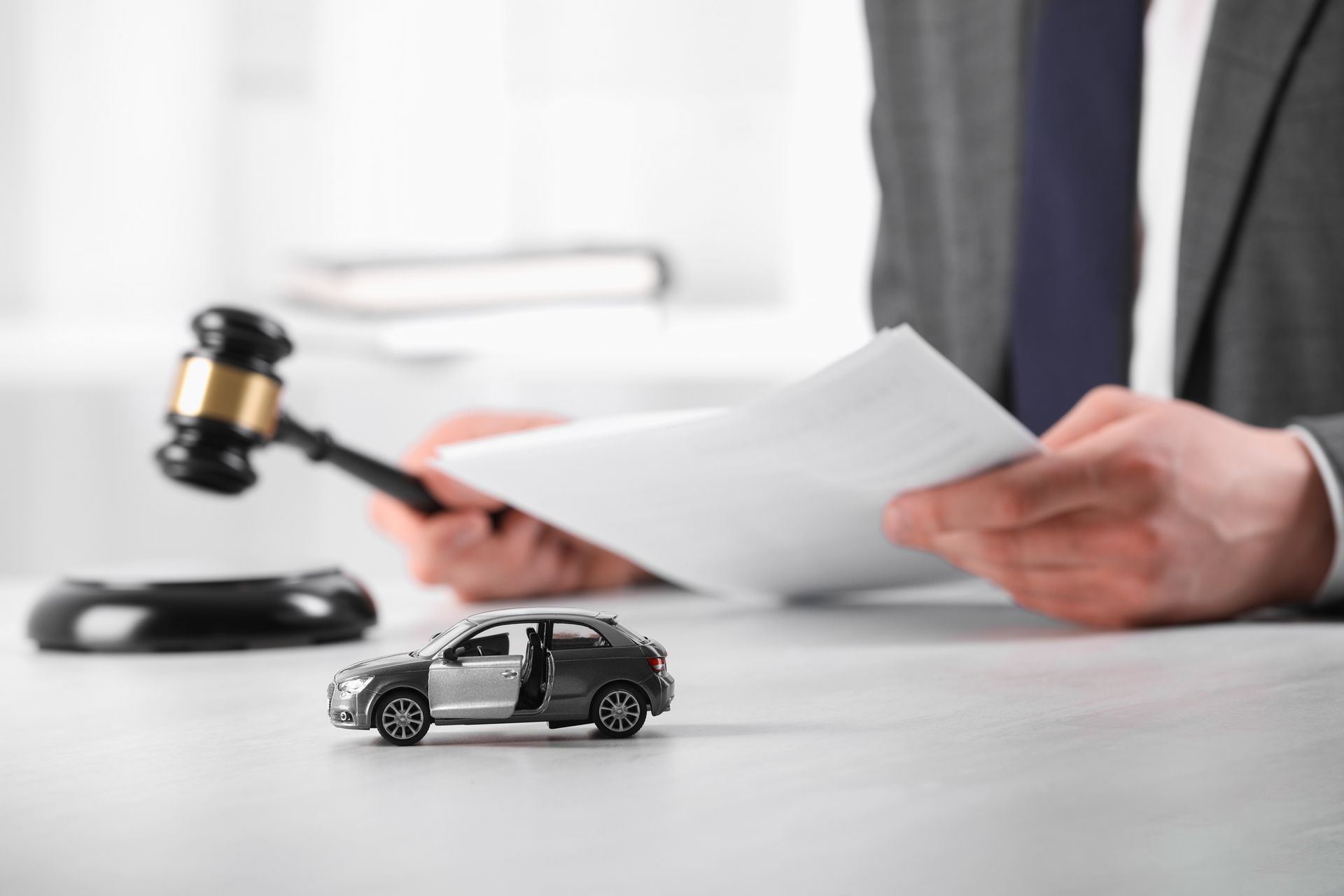 A toy car sits on a desk in front of a person holding papers and a gavel, suggesting a car auction.