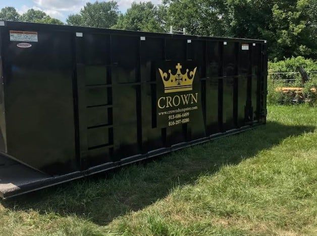 A black dumpster with a crown on it is sitting in the grass.