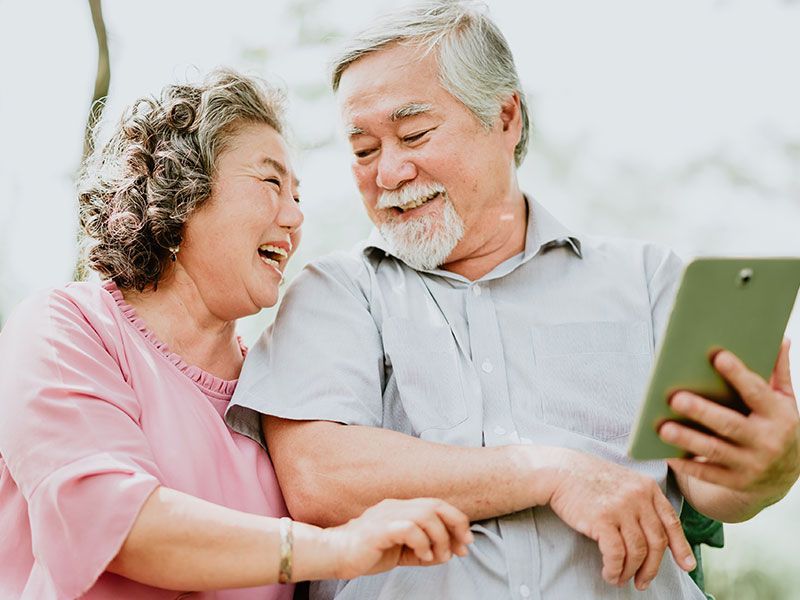 Two people laughing together while looking at a tablet, one holding it up in a bright outdoor setting