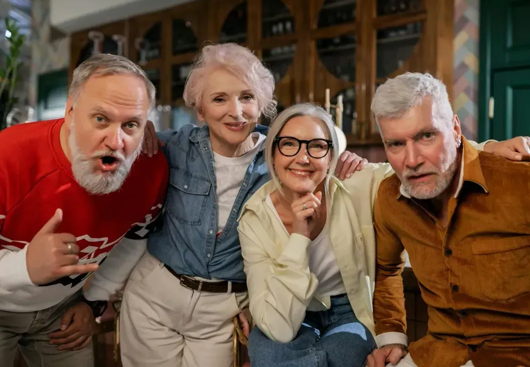 Four smiling adults lean toward the camera in a cozy indoor setting.