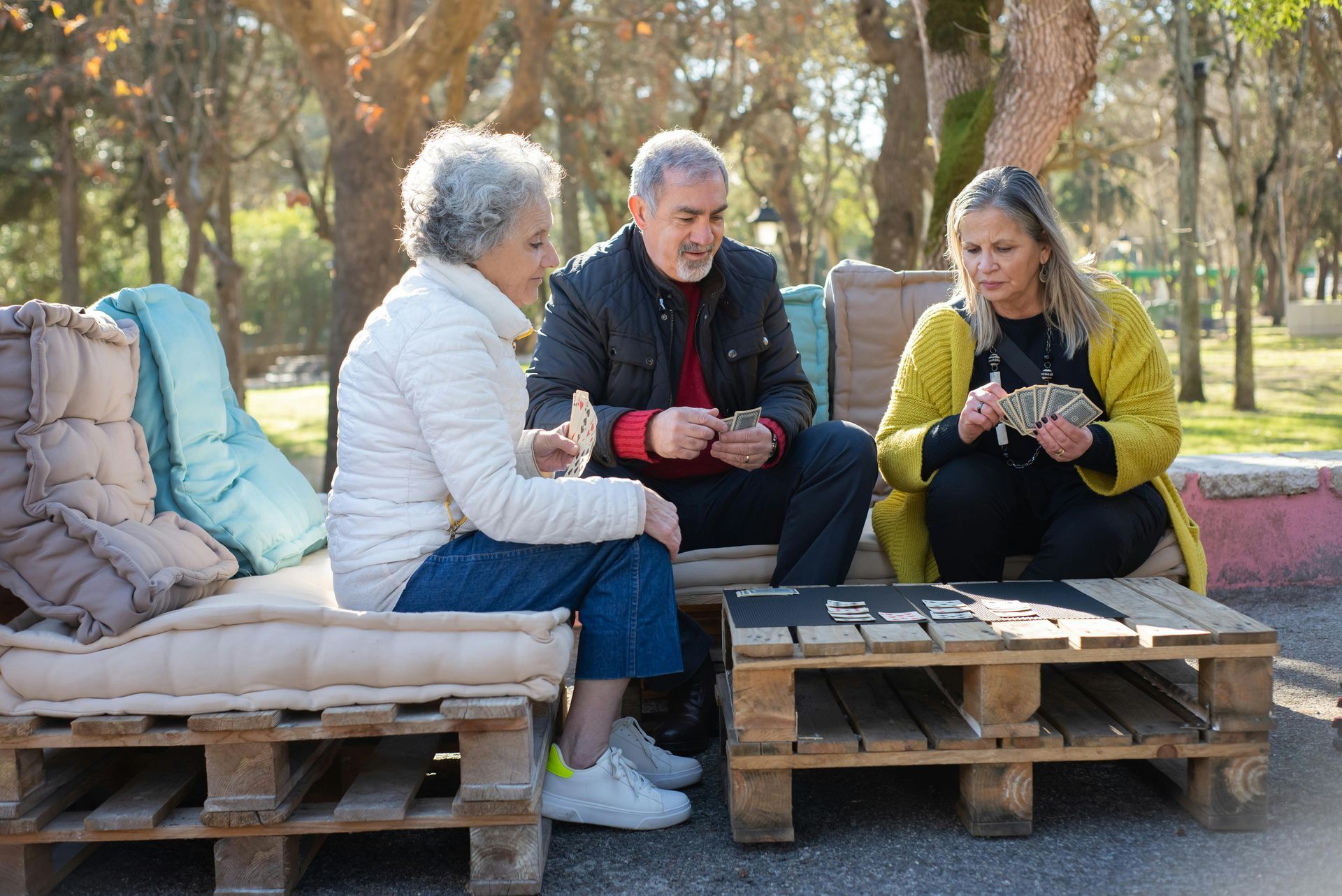 Three people sit on pallet benches outdoors, chatting around a small table in a wooded park.