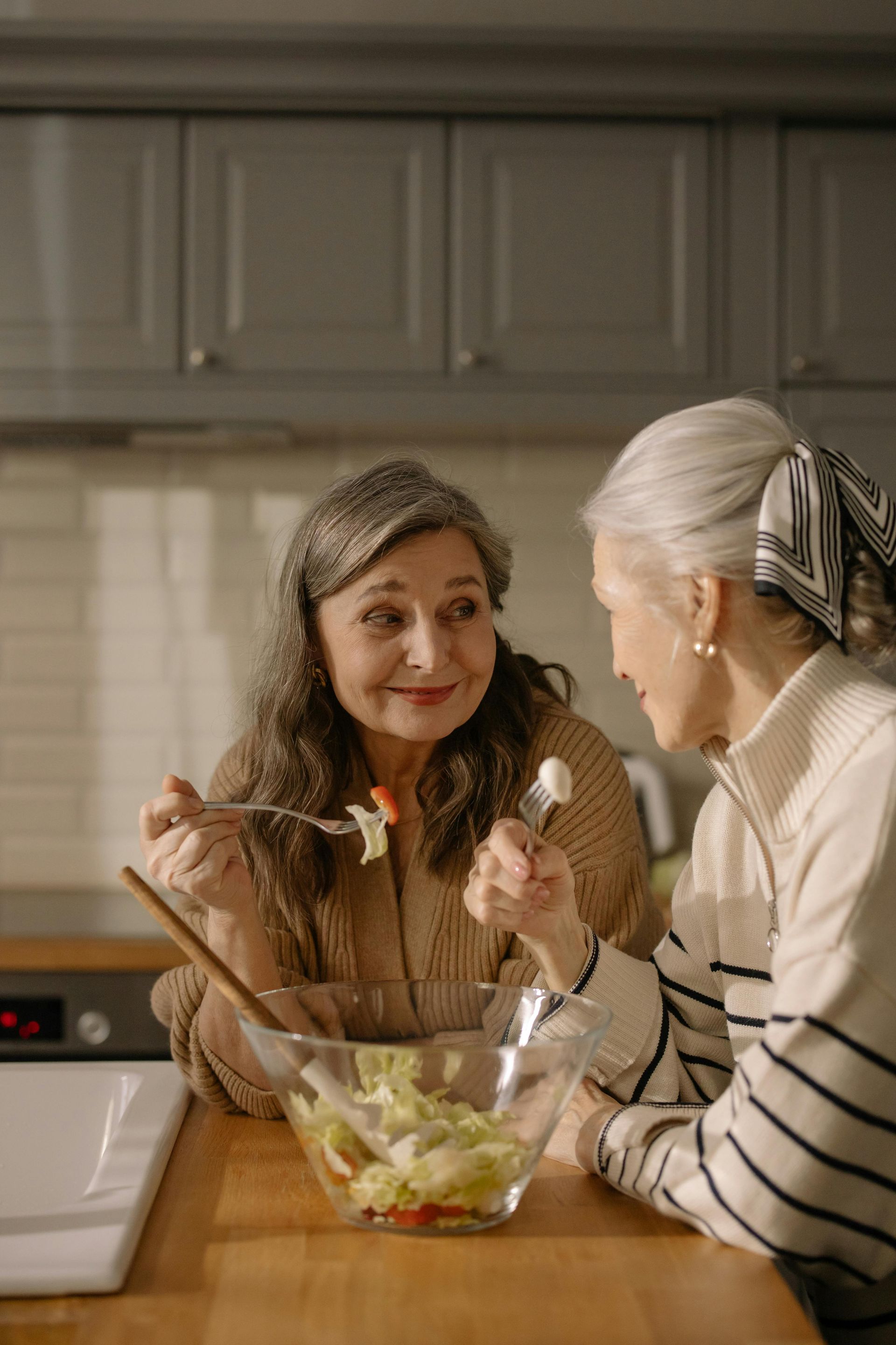 Two people talking over salad in a kitchen, with one holding a fork and a bowl on the counter.
