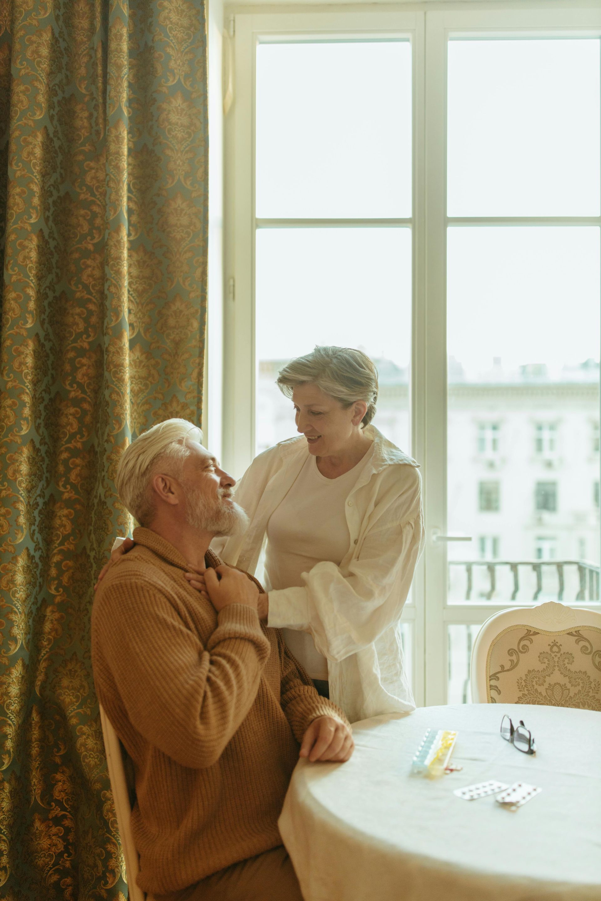 Two people by a bright window, one standing beside a seated person in a cozy room.