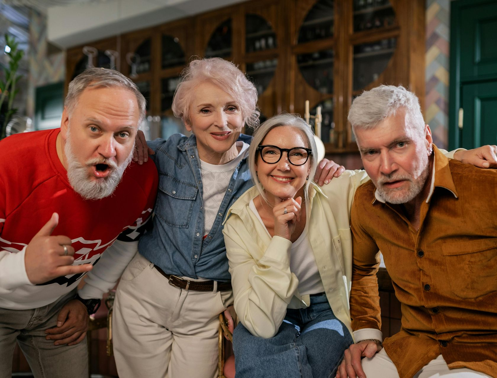 Four smiling adults lean toward the camera in a cozy indoor setting.