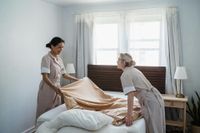 Two women making a bed in a bright bedroom with white curtains and pillows