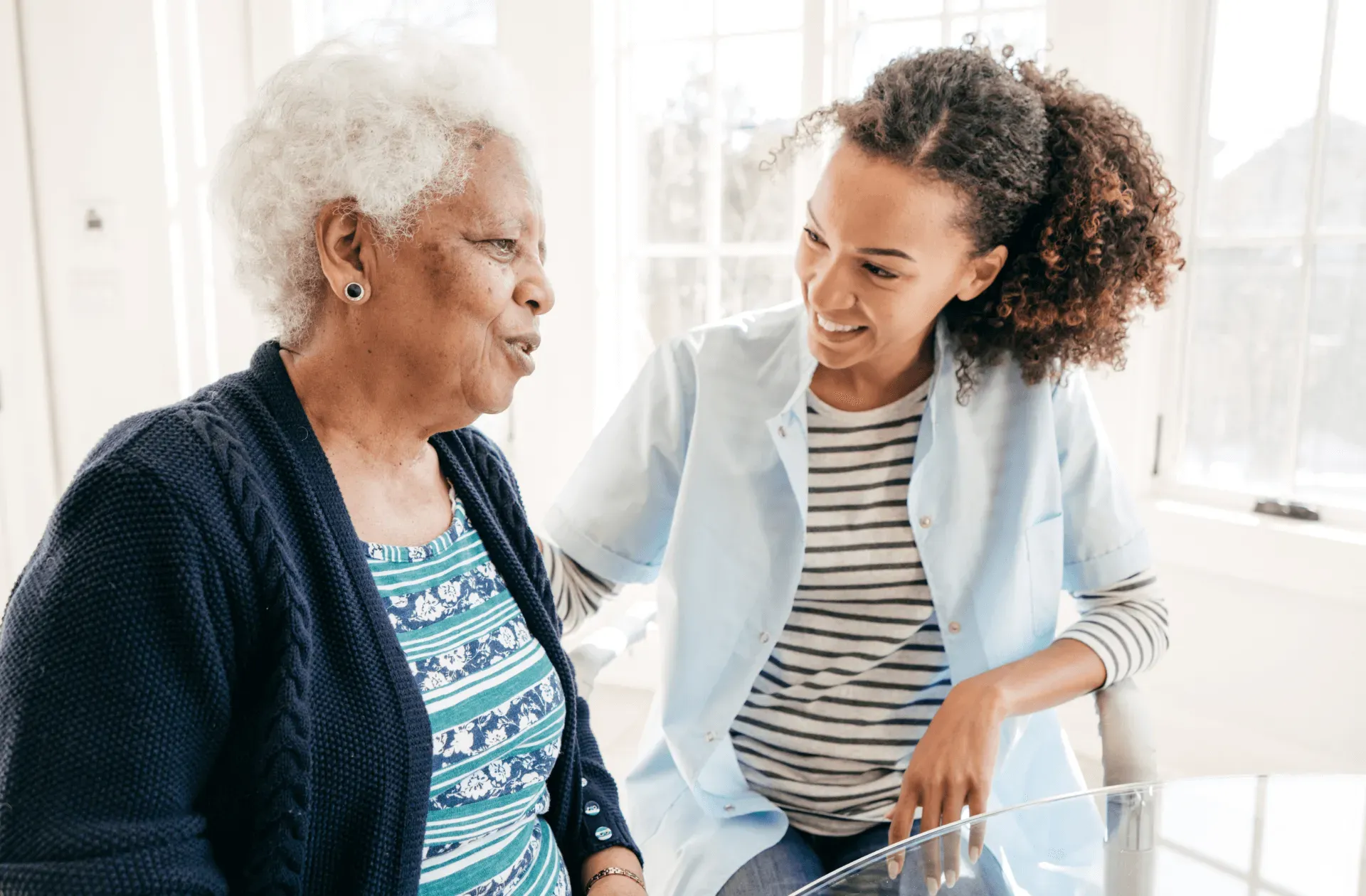 Two women talking in a bright kitchen, one seated and smiling while the other leans in beside her.