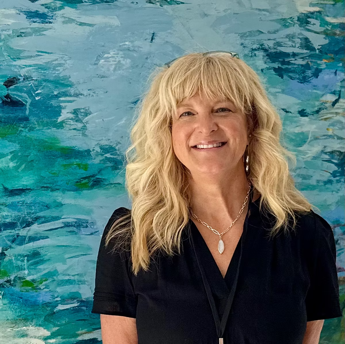 Blonde woman smiling, wearing black top, necklace; standing in front of abstract blue and turquoise painting.