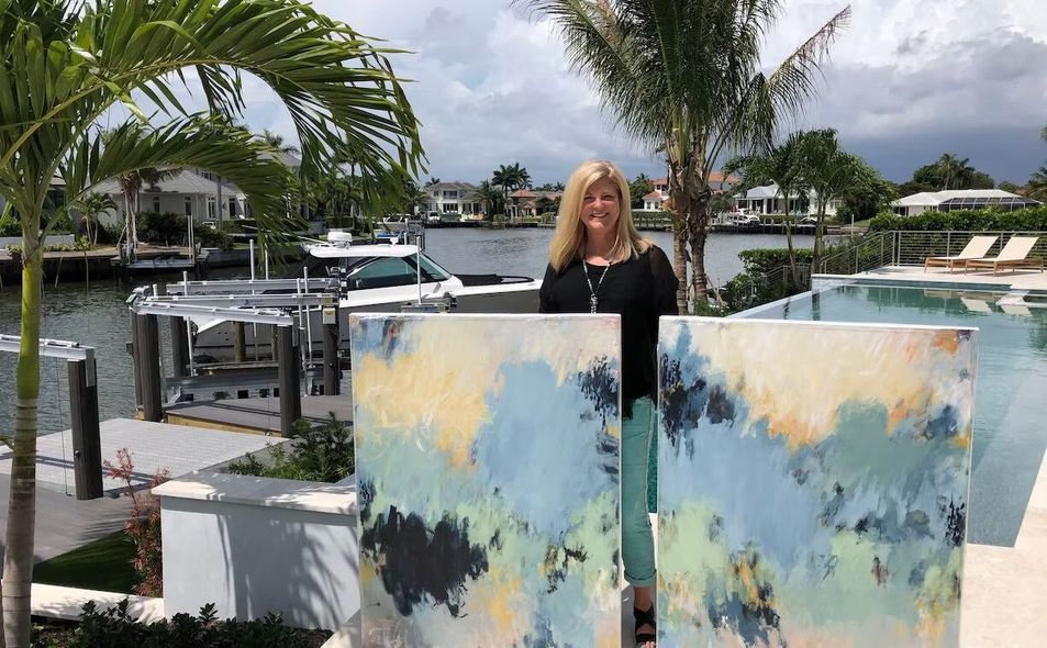 Woman stands between two abstract paintings outdoors near water and palm trees.