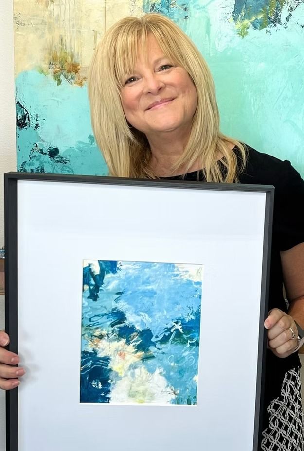 Woman holding framed abstract art with blues and whites; setting is a gallery or studio.