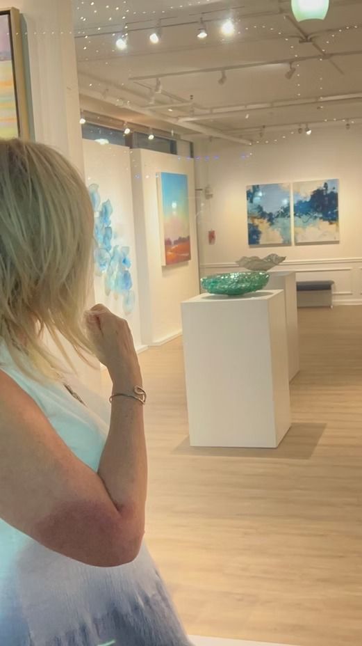 Woman viewing art in a gallery. White walls, wood floor, abstract paintings, and a sculpture on a pedestal.