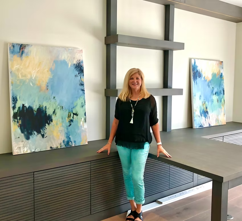Woman stands near abstract paintings and dark shelving in a modern room. She wears turquoise pants, black top.