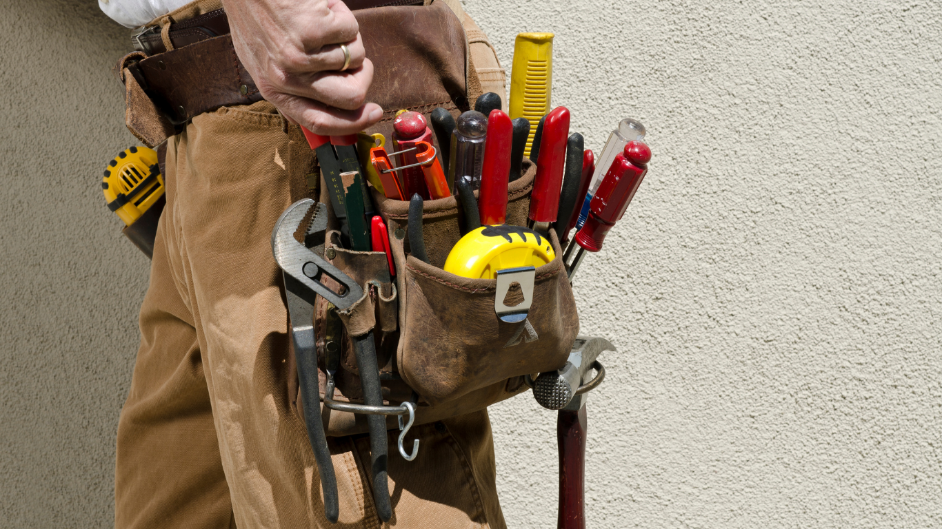 Worker wearing a tool belt filled with screwdrivers and pliers against a plain wall