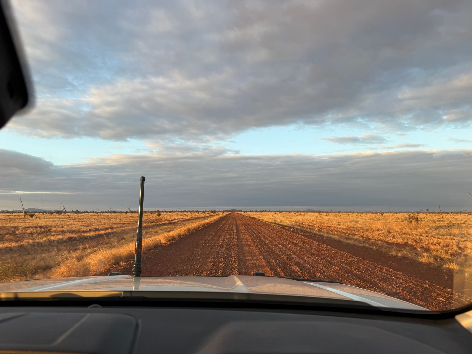 Car driving down a long dirt road through a dry, grassy landscape under a cloudy sky.