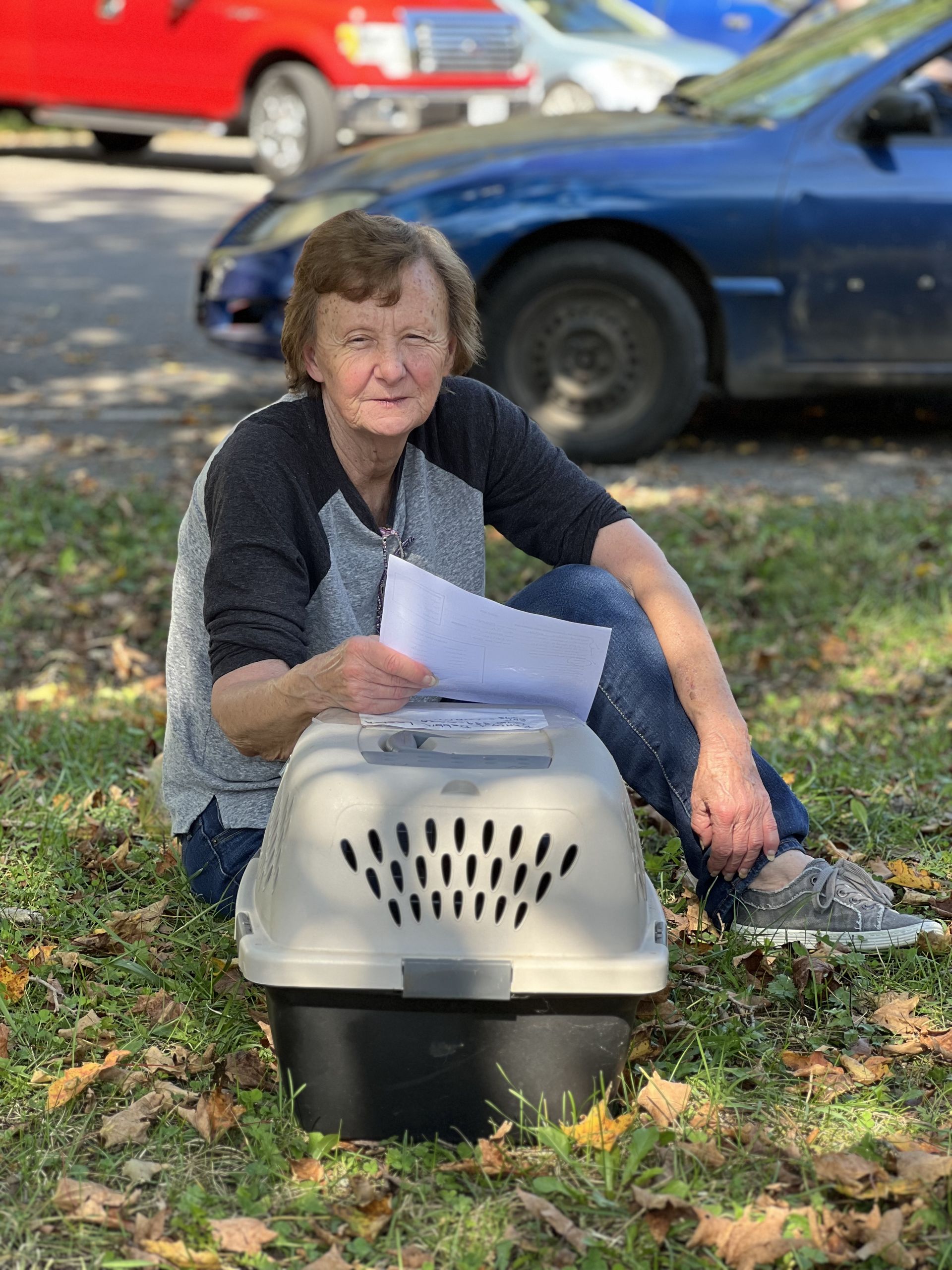 A woman with a cat in a travel carrier sitting on the ground with papers in her hand.