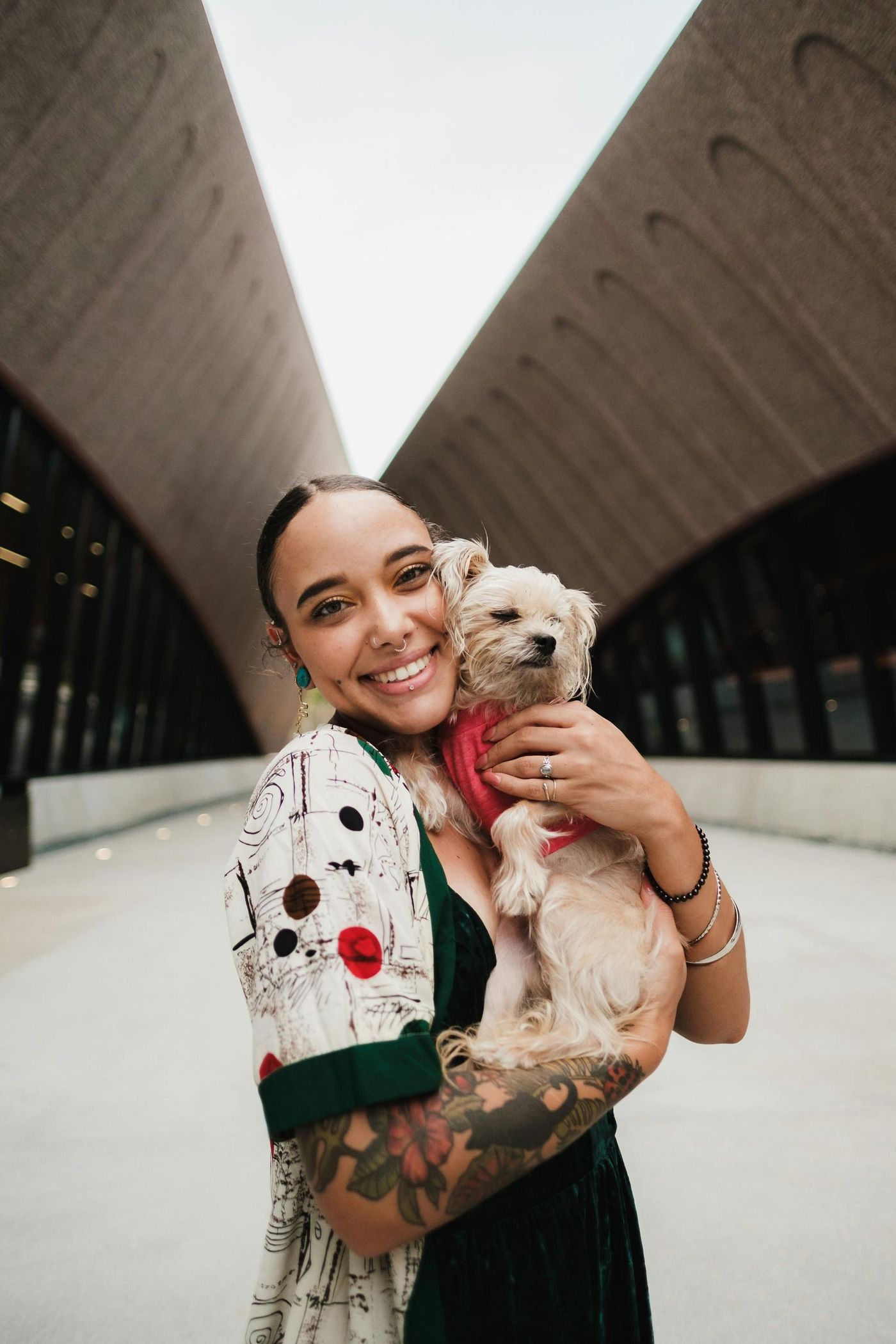 Person smiling and holding a small dog in a modern outdoor walkway between tall buildings.