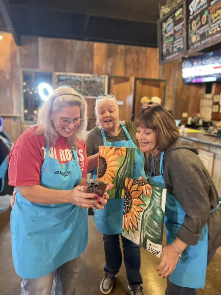 Three people in blue aprons smile and hold a drink in a cozy restaurant.