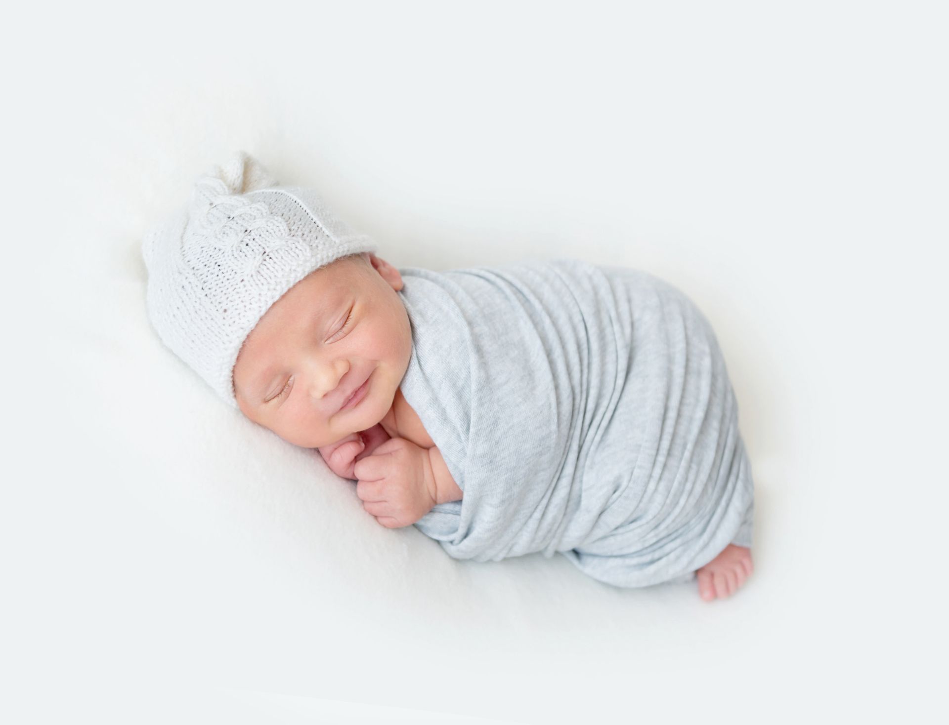 A newborn baby is wrapped in a light gray blanket and wearing a white hat.