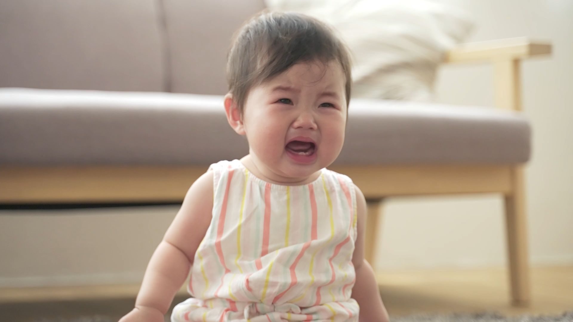 A baby girl is crying while sitting on the floor in front of a couch.