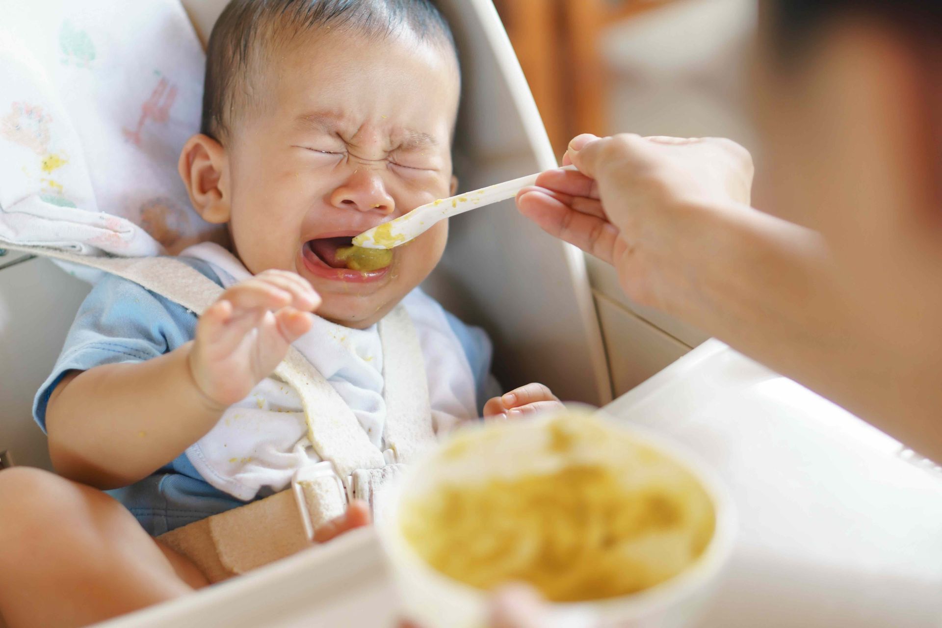 A baby is crying while being fed with a spoon.