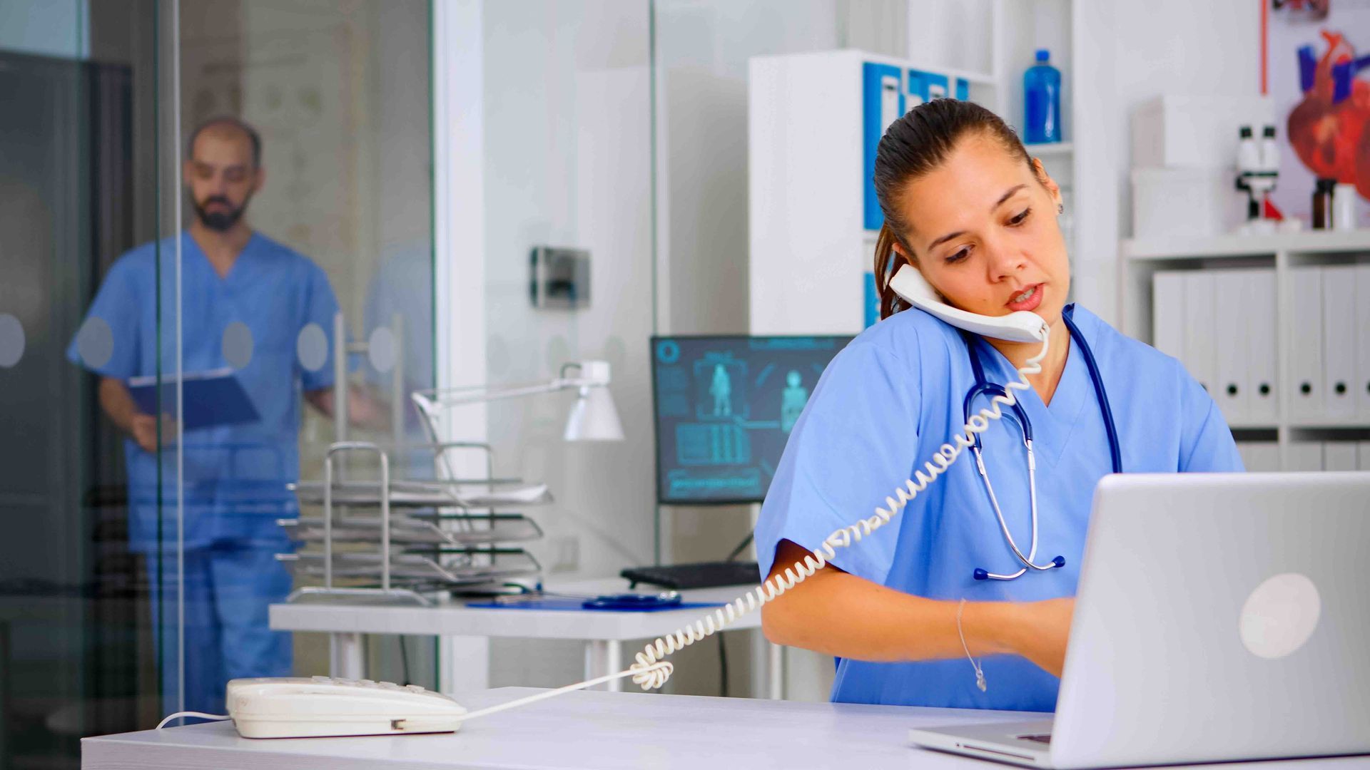 a female doctor is sitting at a desk talking on the phone while using a laptop.