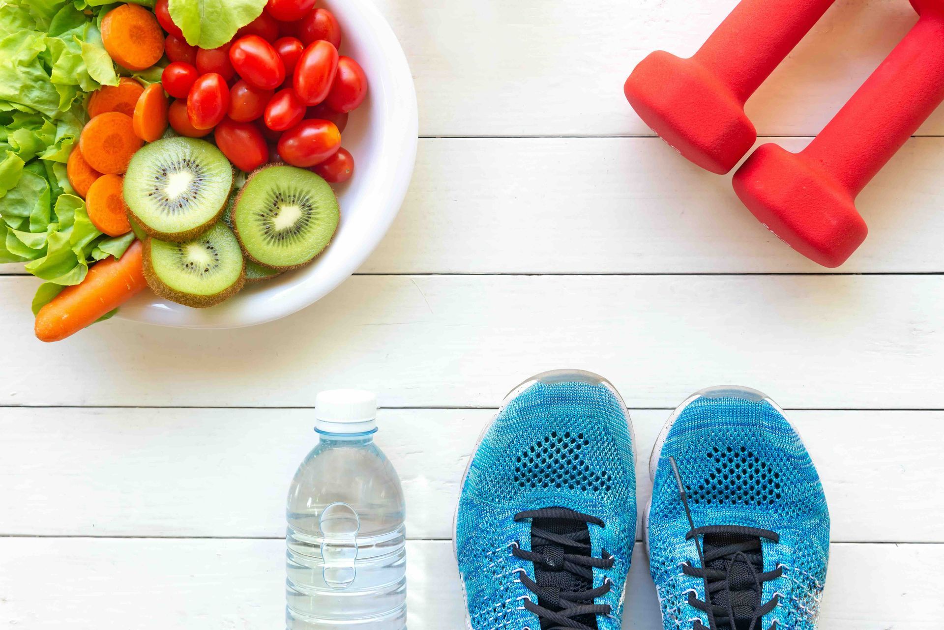 Bowl of salad and fruits next to dumbbells, water bottle, and blue sneakers on a white wood surface.