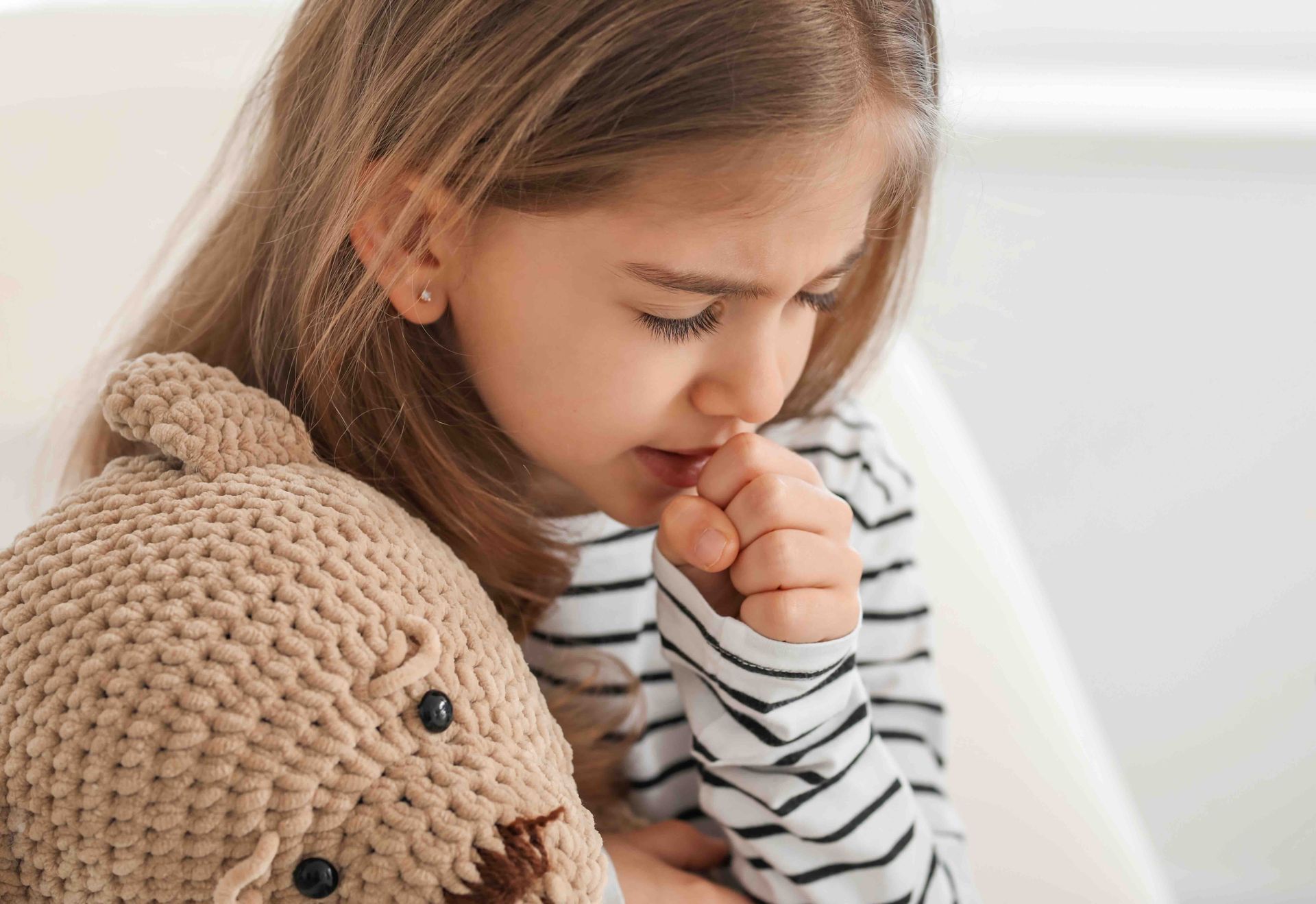 A little girl is coughing while holding a teddy bear.