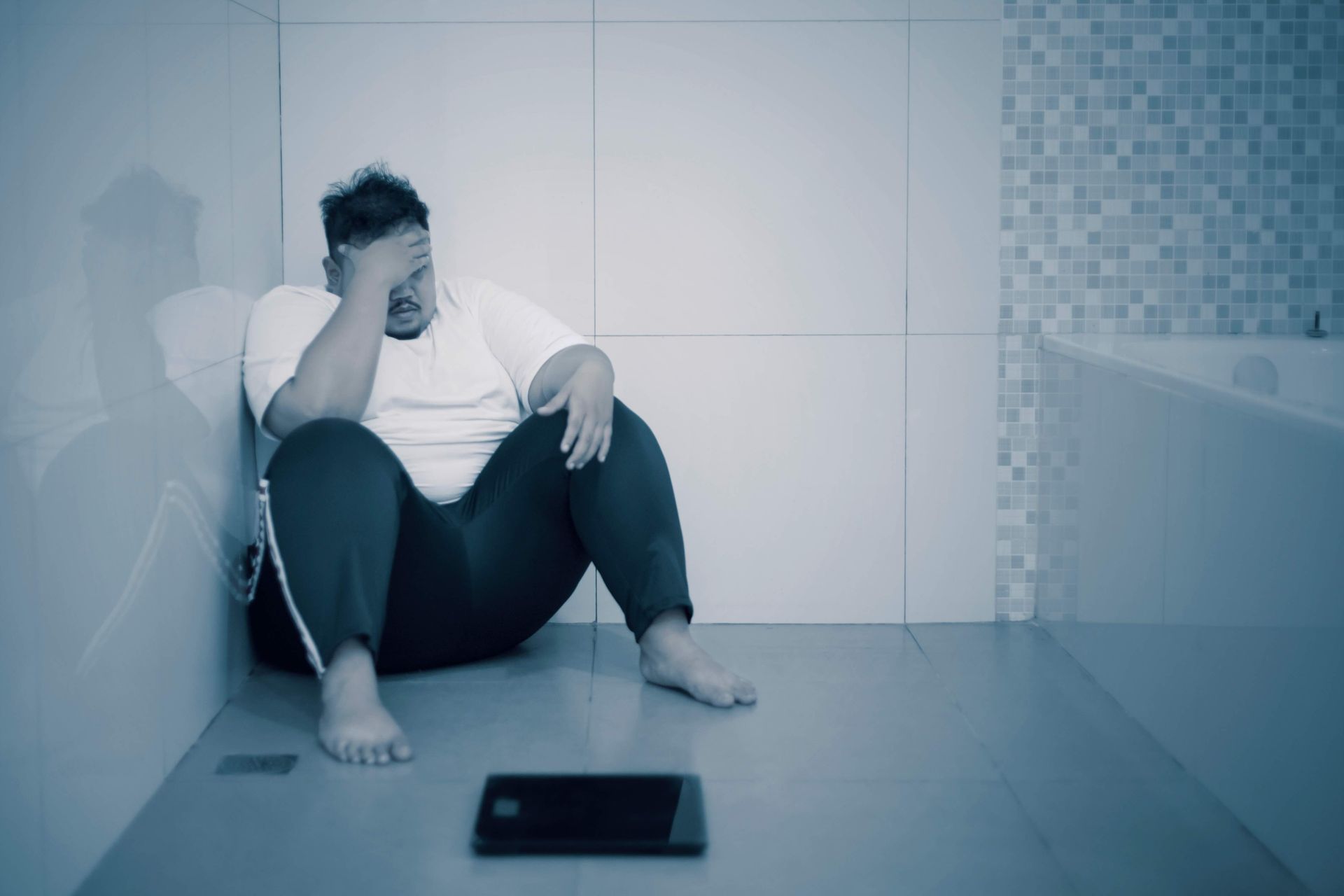 A man is sitting on the floor in a bathroom next to a scale.