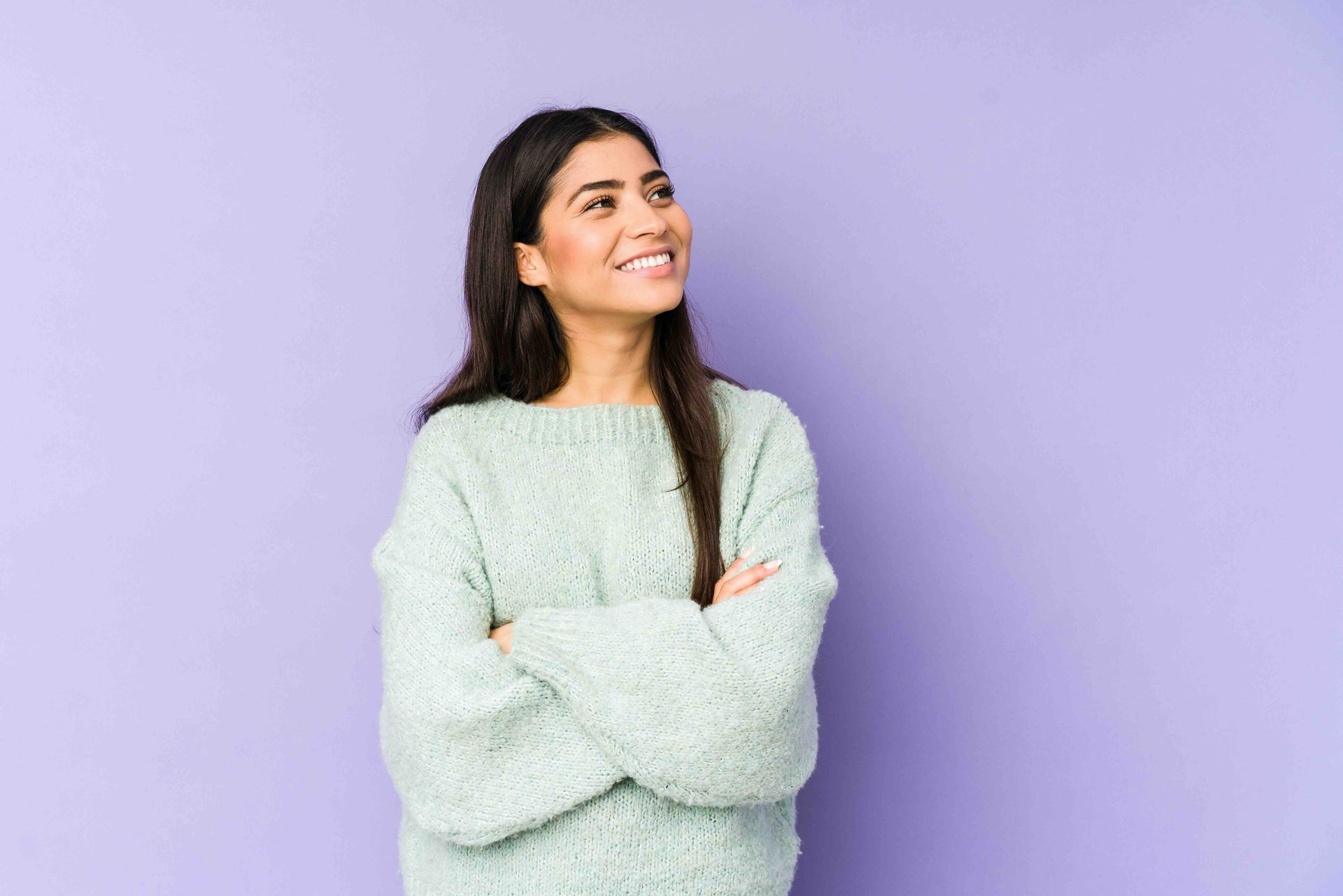 A person with long dark hair wears a light blue fuzzy sweater, smiling with arms crossed against a solid purple background.