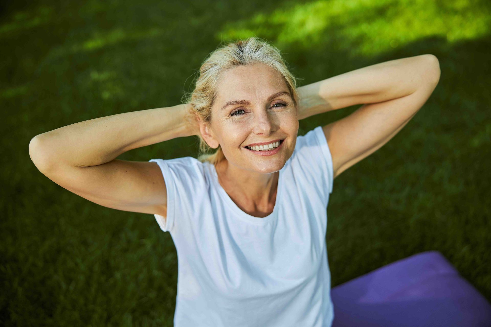 A smiling person in a white t-shirt rests with their hands behind their head while sitting on a mat outdoors on the grass.