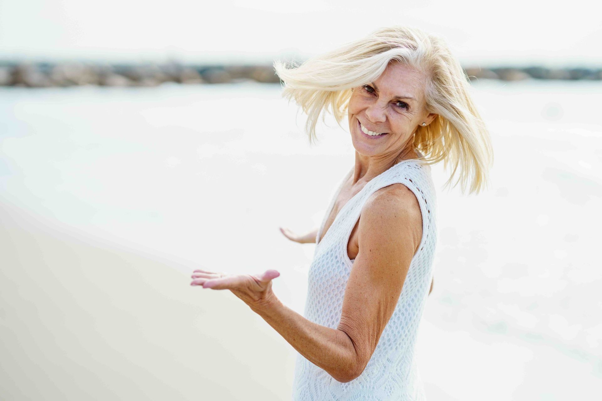 A person in a white dress stands on a beach, smiling with their hair blowing in the wind and hand extended outward.