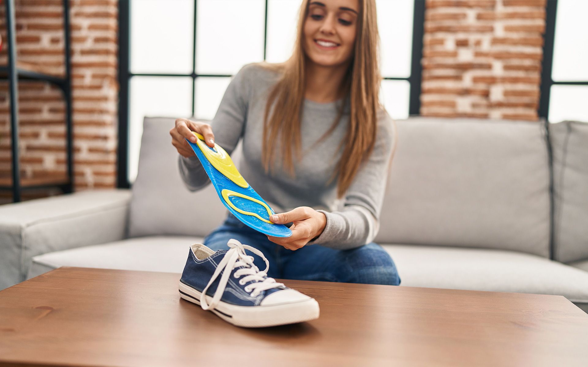 Woman holding a blue and yellow insole, about to put it in a blue sneaker on a table. Smiling, indoors.
