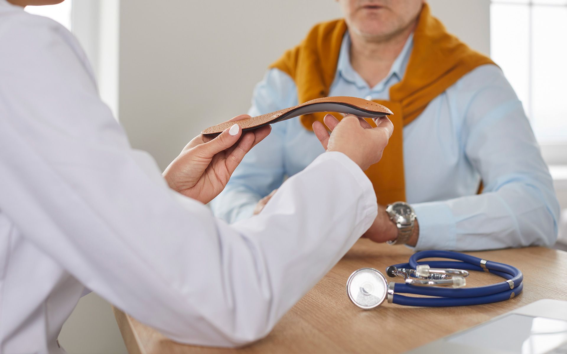 Doctor shows patient a shoe insole in office; stethoscope on desk.