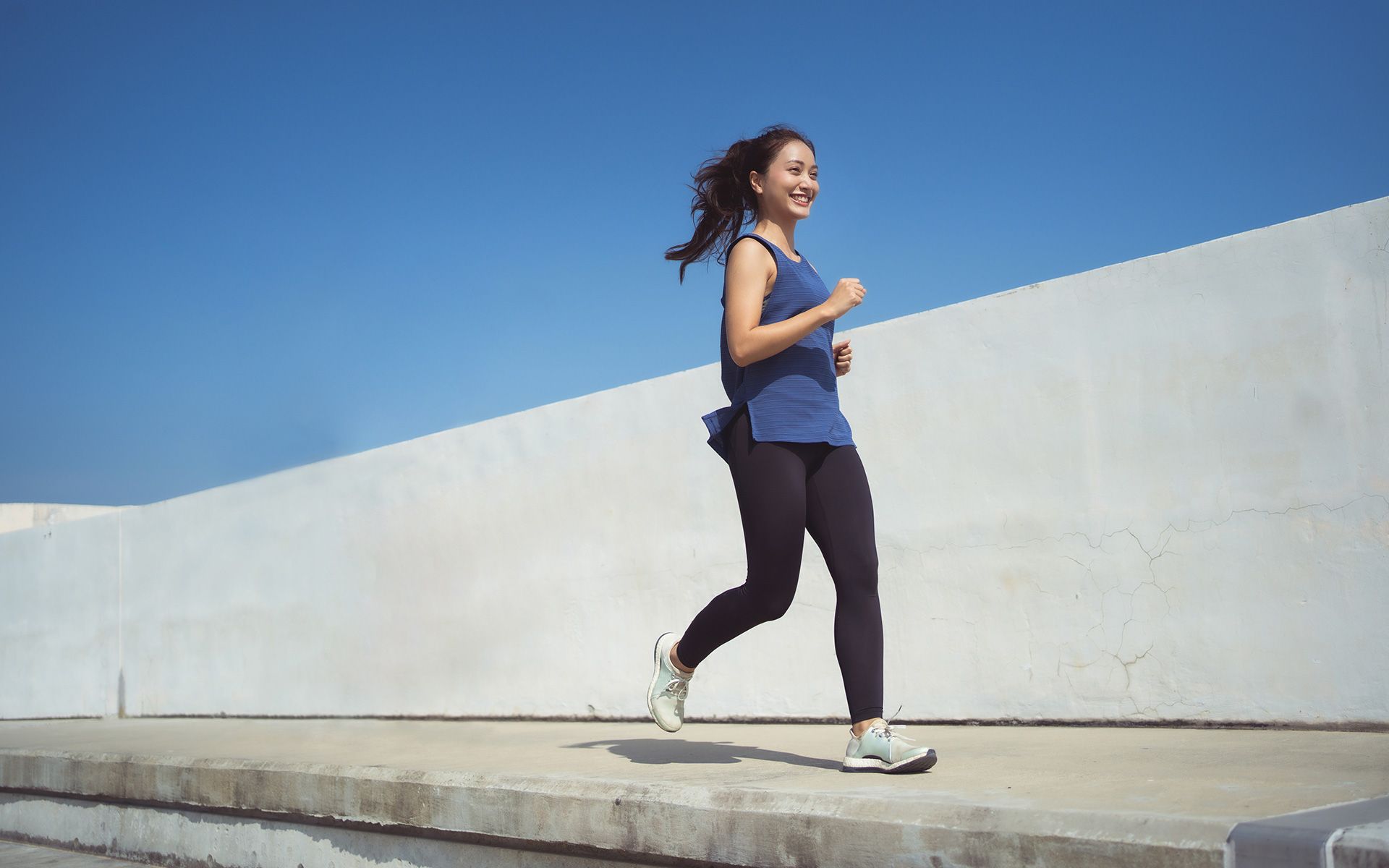 Woman smiling and jogging on a concrete ledge against a blue sky, wearing a blue tank top and black leggings.